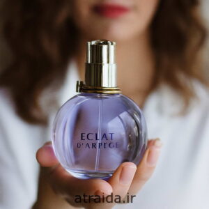 تصویر عطر اکلت زنانه Éclat در دست یک زن حس لوکس و زنانه‌ای به تصویر بخشیده است. در پایین برچسب "atraida.ir" هویت برند عطر آیدا ( امیر عطری ) آمده است.